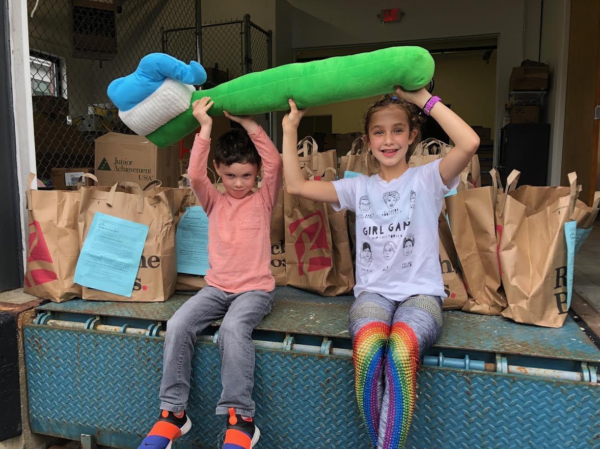 A young boy and girl sit on a loading dock holding Hope and Comfort's iconic stuffed toothbrush over their heads with donation bags filled with toiletries behind them.